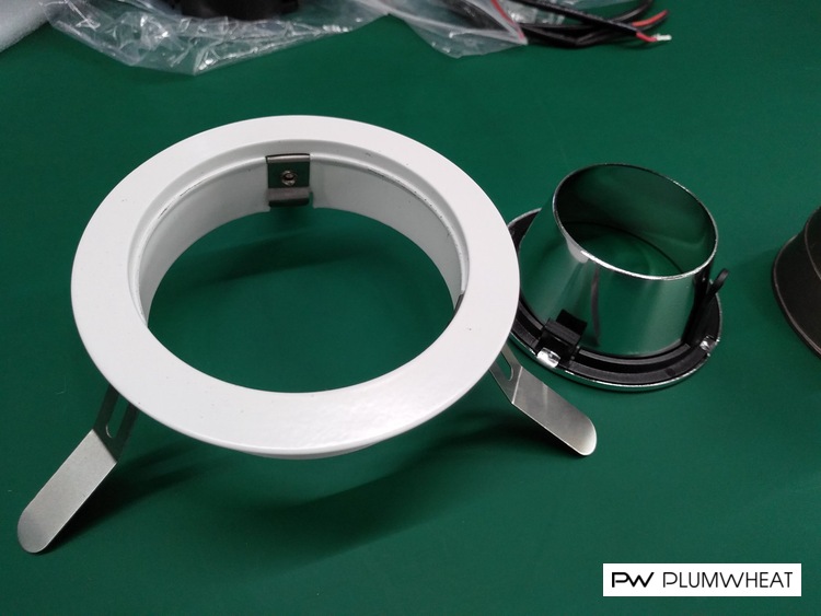 White downlight mounting frame beside a reflective metal component on a green inspection table